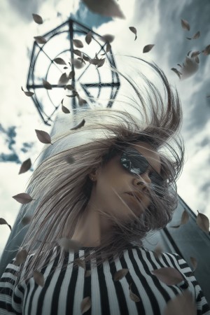 Young blonde woman wearing aviator style sunglasses and a black and white striped shirt her hair swirling in the wind in front of a tower with autumn leaves flying around